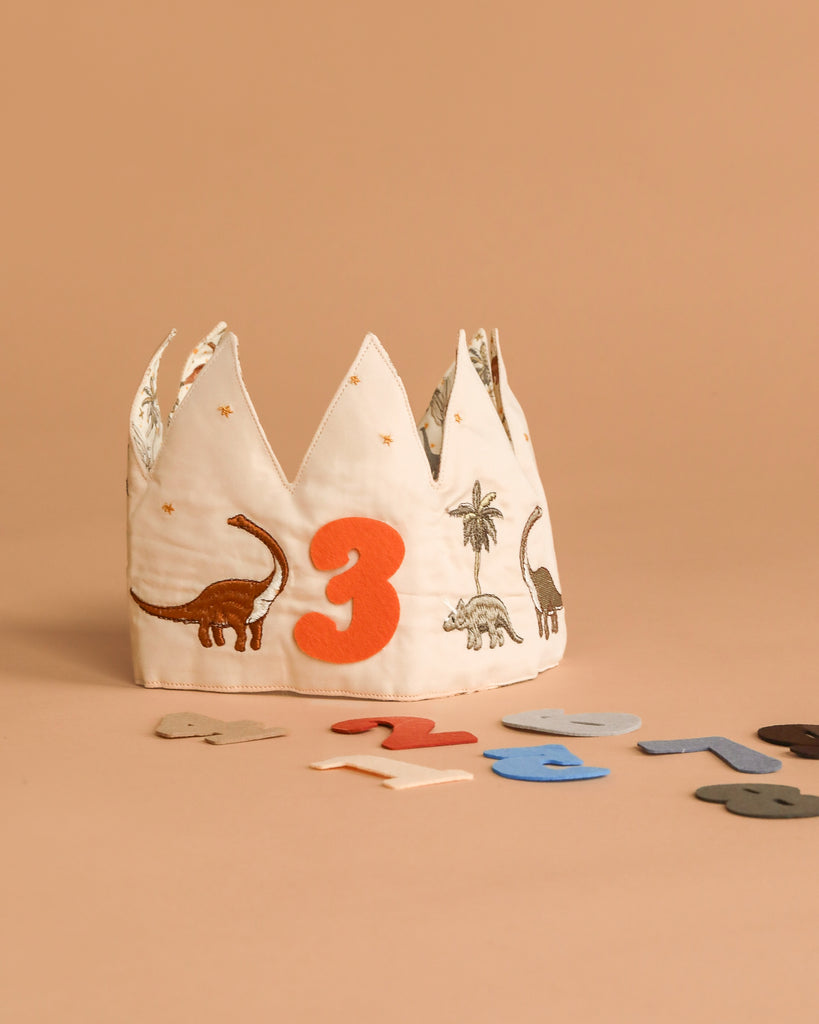 The Konges Sloejd Birthday Crown - Dino features embroidered dinosaurs and a bold orange number 3, with colorful felt numbers. It's a fun, adjustable birthday accessory, shown on a beige surface and background.