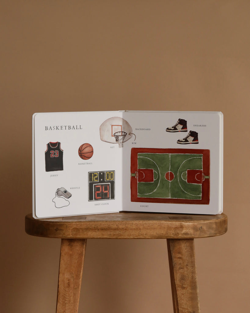 Our Big Book of Sports: A Collection of 100+ Sports Words sits open on a wooden stool, showing basketball items like jersey, ball, hoop, and court—all labeled to help preschoolers build vocabulary. The background is beige.