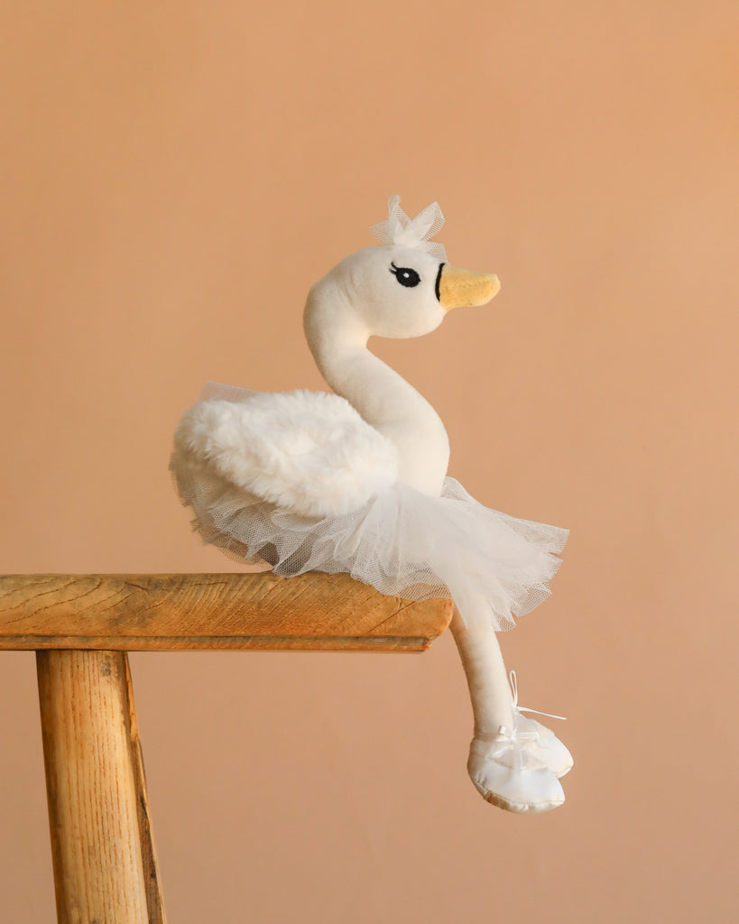 The Bukowski - Odette Ballerina Swan Set features a plush swan in a tulle tutu and ballet slippers, perched on a wooden stool against a beige background—a charming Swedish-designed toy perfect for any nursery.