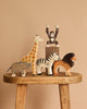 Five animals from the Holztiger Handmade Wooden Safari Animals Set—a giraffe, monkey, elephant, zebra, and lion—are arranged on a wooden stool with a beige background. The monkey stands raised on a small block for added playful charm.