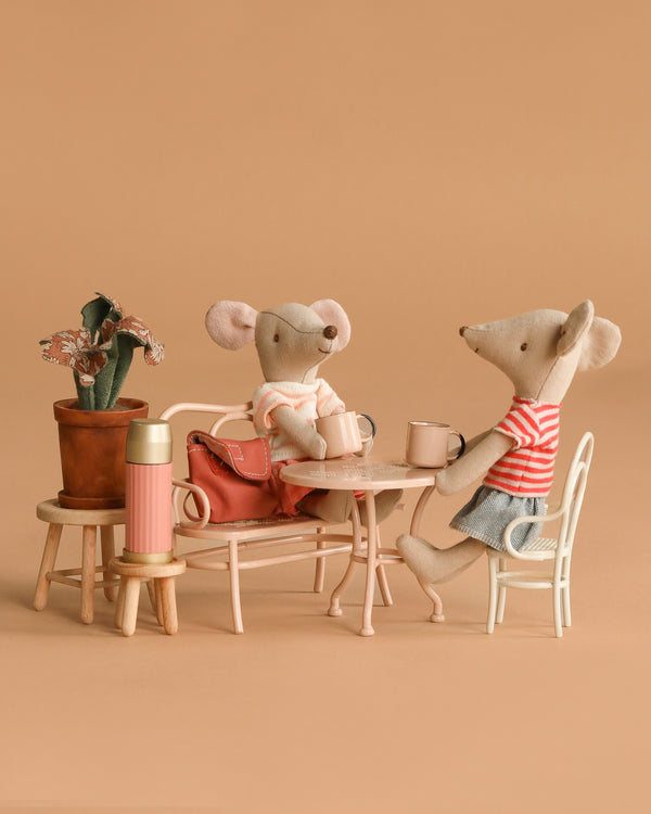The Maileg Outdoor Tea Set features two plush mice, including a big sister mouse in a scarf, sitting at a beige table and bench with mugs. A pink thermos, potted plant, and red bag complete the charming cozy setup.