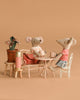The Maileg Outdoor Tea Set features two plush mice, including a big sister mouse in a scarf, sitting at a beige table and bench with mugs. A pink thermos, potted plant, and red bag complete the charming cozy setup.