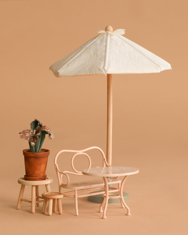 A cute display of the Maileg Patio Set—including a beige parasol, pale pink metal chair and table, plus a potted cactus on a wooden stool—styled against a solid light brown background.