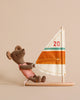 A Maileg Little Sister Surfer Mouse wearing a pink outfit is sitting on a wooden platform, holding onto an orange, white, and brown striped sail with the number 20 on it. This adorable surfing toy features magnetic hands for better grip. The background is beige.