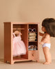 A Maileg Wardrobe, Large - Dark Powder (compatible with Minikane Dolls), stands in a light beige room, featuring a doll in white underwear, a pink dress on a hanger, bunny and teddy clothes, folded outfits, sunglasses, and a drawer.