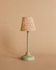 A charming small table lamp from the Maileg Movie Night Set, featuring a pale green base and a patterned lampshade with red and gray accents, set against a beige background—ideal for accessorizing your Maileg dollhouse or enhancing your Maileg furniture collection.