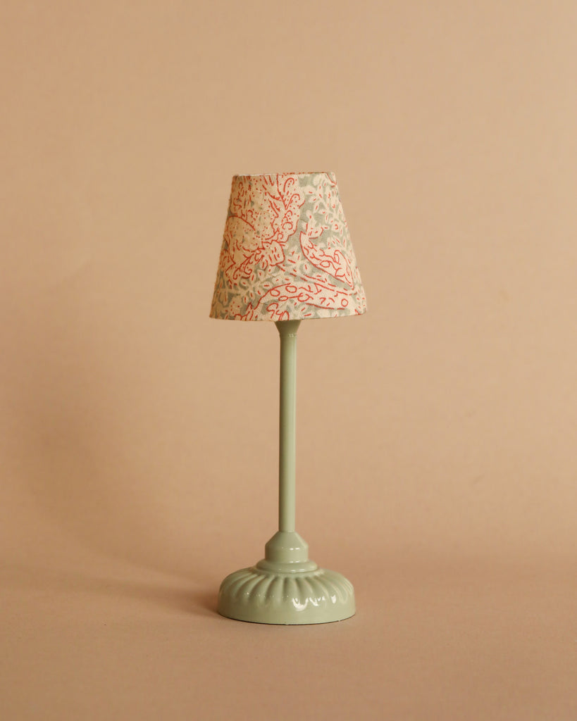 A charming small table lamp from the Maileg Movie Night Set, featuring a pale green base and a patterned lampshade with red and gray accents, set against a beige background—ideal for accessorizing your Maileg dollhouse or enhancing your Maileg furniture collection.