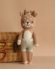 Cuddle + Kind Elliott the Fawn, a hand-knit plush deer with beige fur, white accents, antlers, and light green shorts, sits upright before a woven wicker basket on a neutral beige background.