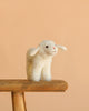 The Steiff Anni Lamb, a white plush toy, stands on a rustic wooden stool against a light peach background—an adorable addition to any Easter basket.