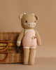 Cuddle + Kind Ava the Cow - Powder Pink is a hand-knit plush doll in a pink outfit, standing upright beside a wicker basket with a metal clasp, set against a plain beige background.