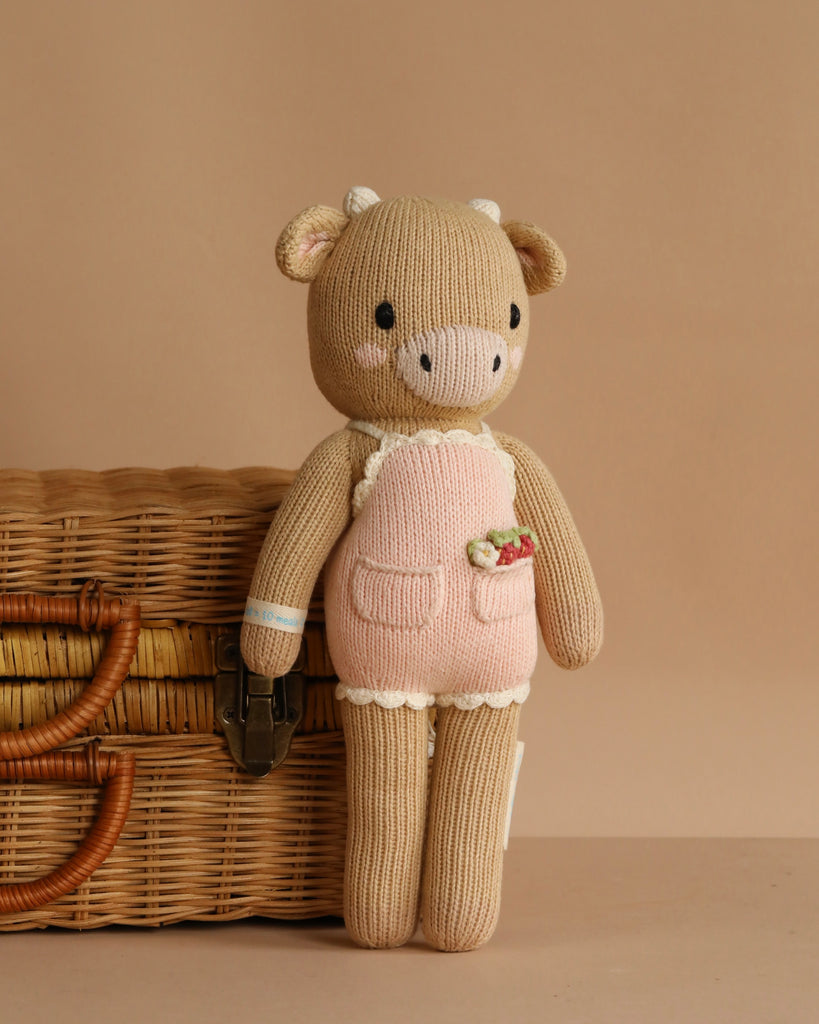 Cuddle + Kind Ava the Cow - Powder Pink is a hand-knit plush doll in a pink outfit, standing upright beside a wicker basket with a metal clasp, set against a plain beige background.