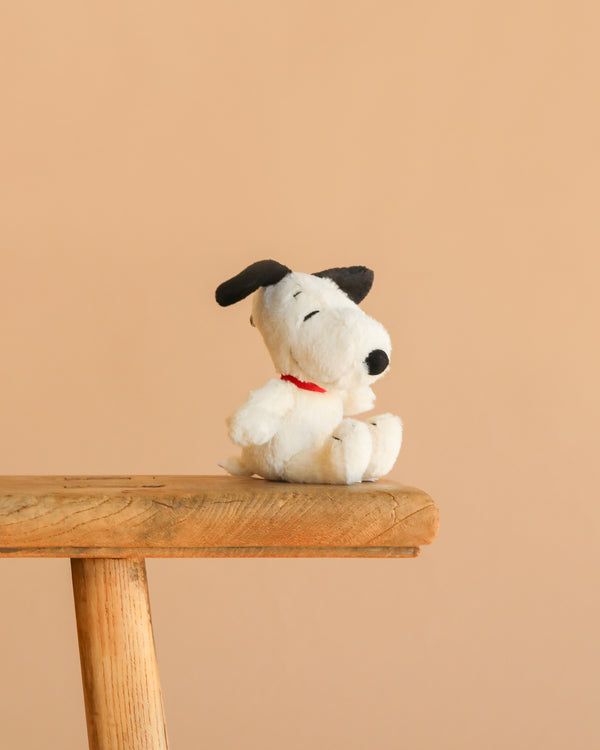 The Steiff Snoopy Keyring, featuring a small Snoopy plush with a red collar, sits on the edge of a wooden stool against a beige background—perfect as an accessory for your backpack.