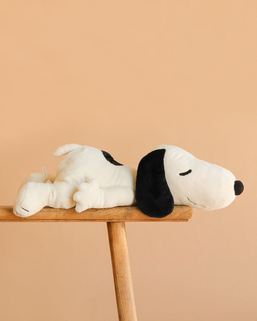 The Steiff XL Snoopy Lying Down 16" plush, a large beagle stuffed animal, rests on its stomach atop a wooden bench with a plain beige background.