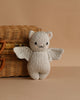 The Cuddle + Kind Baby Limited Edition Grey Bat, a light gray plush toy with bat wings, round ears, and a smiling face made from heirloom-quality Peruvian cotton yarn, sits before a wicker basket on a beige surface.