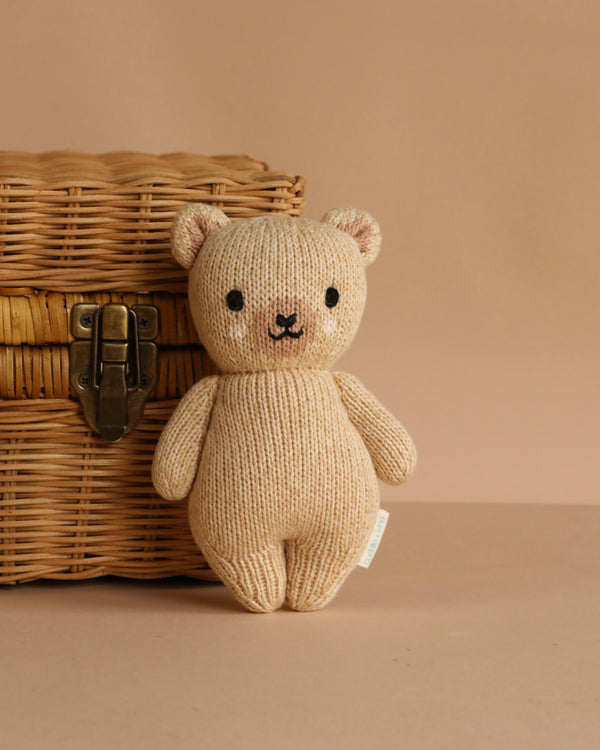 The Cuddle + Kind Baby Honey Bear, a small, beige hand-knit bear made from heirloom-quality Peruvian cotton yarn, sits beside a wicker basket with a metal latch against a plain beige background.