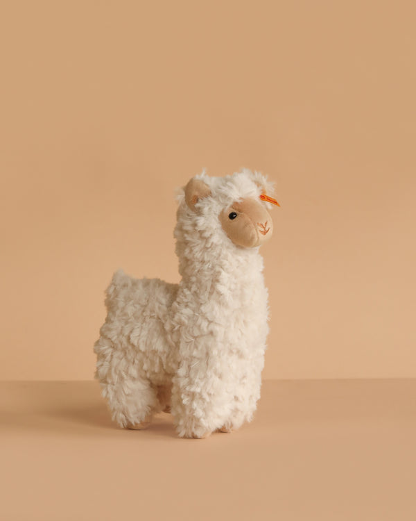 The Steiff Leandro Llama Plush Toy, 7 inches, features soft white fur and an orange bow by its ear. From the Soft Cuddly Friends collection, it stands on a beige surface—an ideal cuddly companion for all ages.