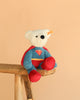 The Steiff Superman Teddy Bear, 12 Inches, features a white bear in a blue and red costume with a red cape, sitting on a wooden stool against a beige background—the perfect superhero plush for any fan.