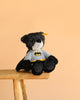 The Steiff Batman Teddy Bear, 12 Inches, dressed in a black and gray superhero costume, sits on a wooden stool against a plain peach-colored background.