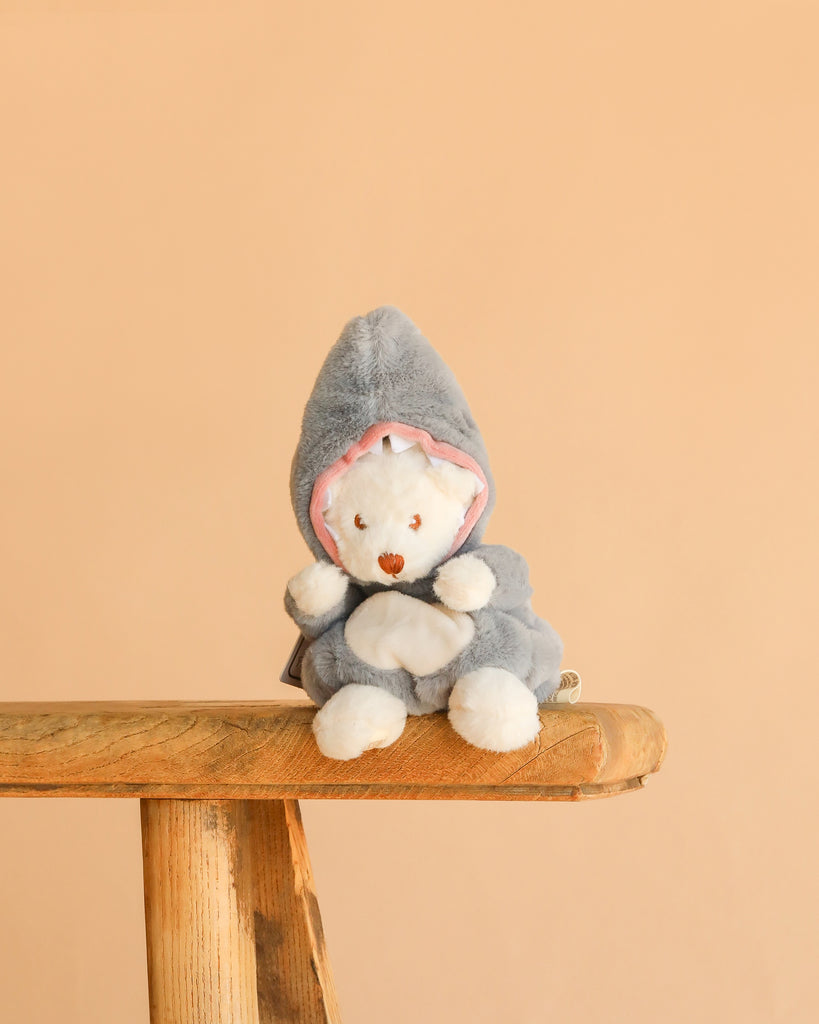 Bukowski - Ziggy Shark is a white teddy bear dressed in a gray shark costume with pink lining, seated on a wooden bench against a beige background—an adorable collectible plush.