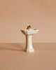 A charming Maileg Suitcasa Furnished Set, featuring a small white pedestal sink with a gold faucet, is displayed against a simple beige background.