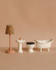 Four miniature bathroom accessories are arranged in a row on a beige surface: a floral lamp, white pedestal sink with gold faucet, Maileg Toilet, Mouse Size - Off White, and a classic clawfoot tub—perfect for your SuitCasa mouse house.