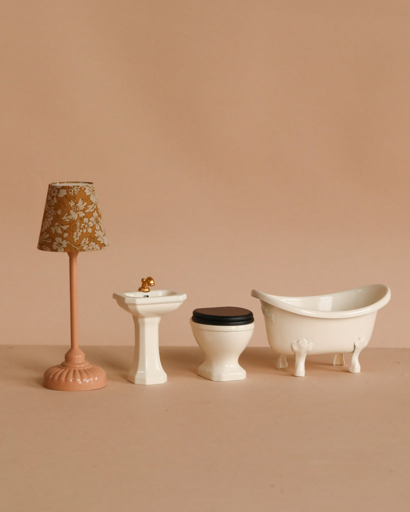 Four miniature bathroom accessories are arranged in a row on a beige surface: a floral lamp, white pedestal sink with gold faucet, Maileg Toilet, Mouse Size - Off White, and a classic clawfoot tub—perfect for your SuitCasa mouse house.