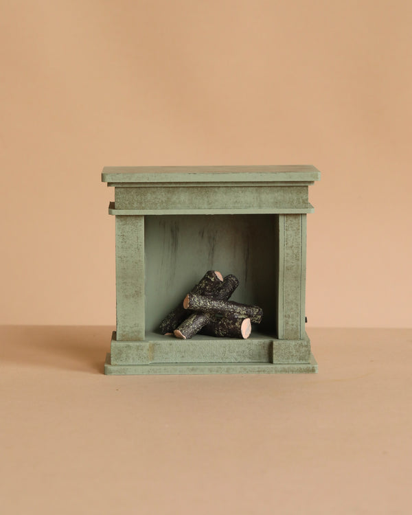 The Maileg Fireplace, Miniature – Dusty Green is a small, green, rectangular wooden fireplace with a vintage finish and stacked logs inside—a charming touch for any miniature home.