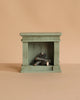 The Maileg Fireplace, Miniature – Dusty Green is a small, green, rectangular wooden fireplace with a vintage finish and stacked logs inside—a charming touch for any miniature home.