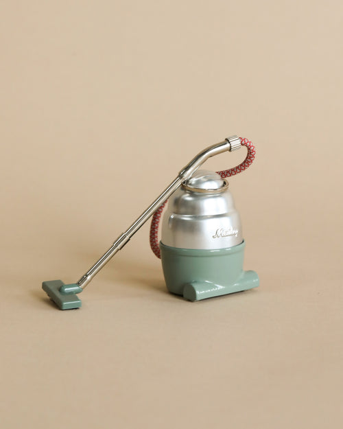 A Maileg Miniature Vacuum Cleaner model in pastel green, displaying "darling" on its front, with a corrugated red hose and a long, thin metal handle, set against a plain
