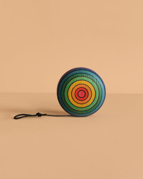 The Mader Wooden Yoyo - Rainbow features handcrafted concentric rainbow circles and a black string, resting on a beige surface. This vibrant, non-toxic toy offers a playful option for kids and collectors alike.