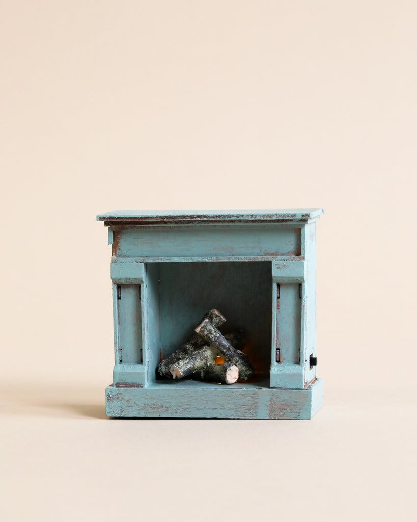 The Maileg Cozy Green Room Set features a small, distressed blue wooden miniature fireplace with faux logs inside—perfect for Maileg mouse furniture or pairing with other mini accessories, set against a plain beige background.