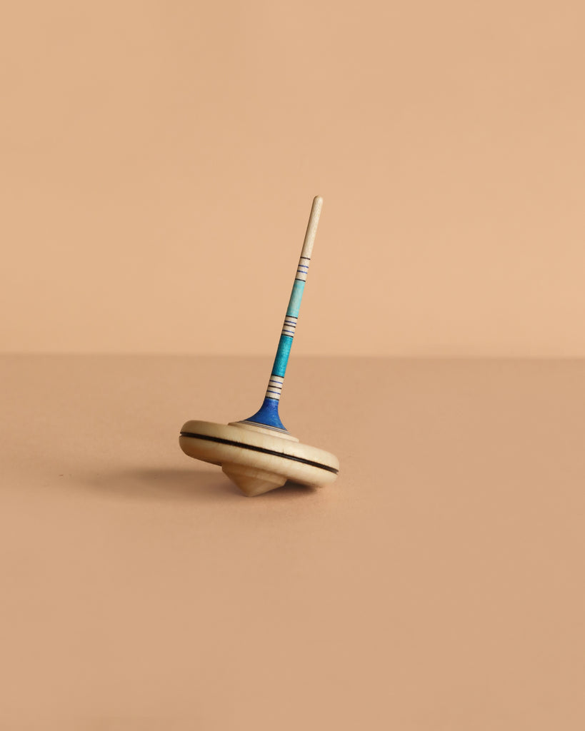 The Mader Spaghetti Spinning Top - Blue, crafted from maple wood with blue and turquoise accents, stands upright on a plain beige surface against a matching background, showcasing its classic charm and safe design.
