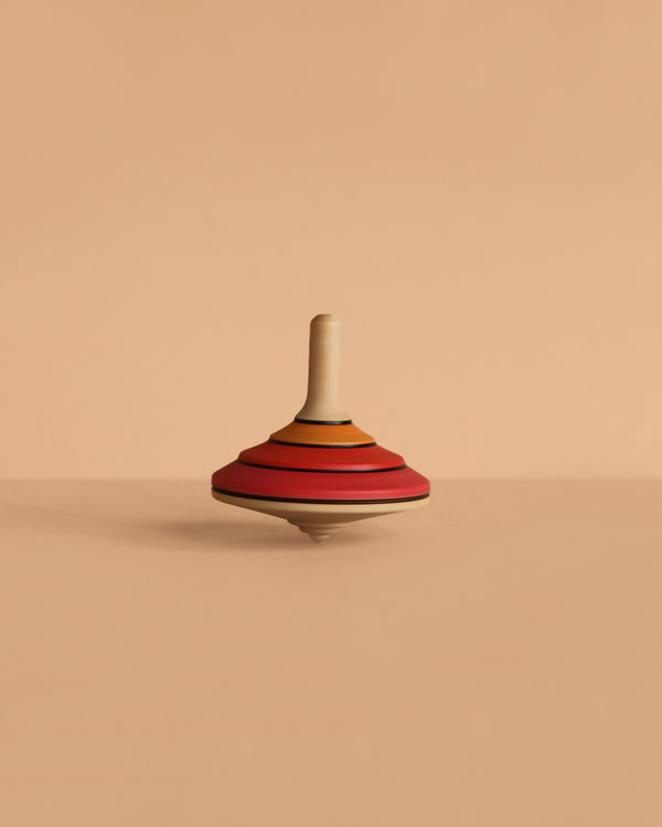 The Mader Tango Spinning Top - Red, featuring red, orange, and tan stripes and crafted from maple wood, stands upright on a smooth beige surface with a matching background.