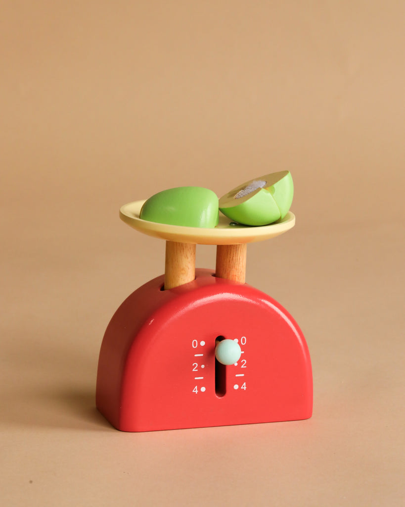 A red Kitchen Weighing Scales toy with a cream tray holds two green apple slice toys, displayed against a plain beige background.