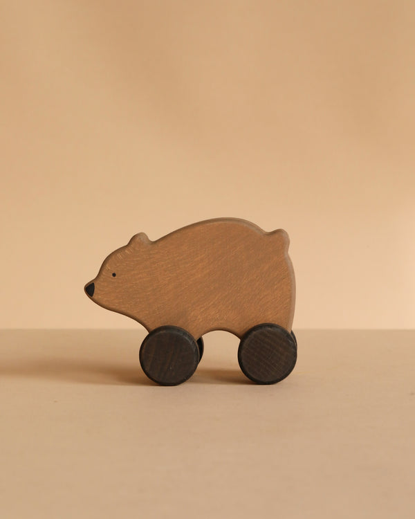 The Sabo Concept Wooden Rolling Brown Bear, featuring simple details and two black wheels for legs, is displayed on a beige surface against a matching beige background.