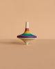 The Mader Large Spinning Top - Rainbow, featuring vibrant stripes, stands upright on a beige surface against a matching beige background.