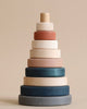 A Wooden Pyramid Stacker - Terracotta featuring rings of varying sizes, painted with non-toxic paint in different muted colors, arranged in order from largest at the bottom to smallest at the top, against a beige background.