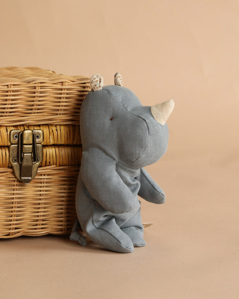 The Maileg Rhino, Small - Dusty Blue, sits beside a closed wicker basket with a metal latch, set against a beige background.