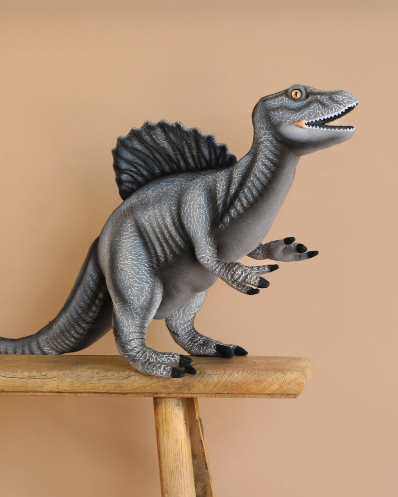 A gray Spinosaurus Dinosaur Stuffed Animal with a large sail on its back stands on a wooden stool against a beige background. It features a textured body, open mouth with teeth, and small forelimbs with claws, highlighting exquisite craftsmanship.