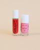 Two bottles of Nailmatic water-based nail polish with cute faces drawn on them, one pink and one red, against a soft peach background.