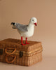 A realistic Seagull Bird Stuffed Animal with a white and gray body and red beak standing on top of a woven wicker basket against a plain light brown background.