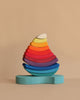 Grimm's Stacking Tower Sailboat features semi-circular wooden blocks, arranged by size and color in a rainbow teardrop shape on a light blue base—perfect for fine motor skill development against a beige background.