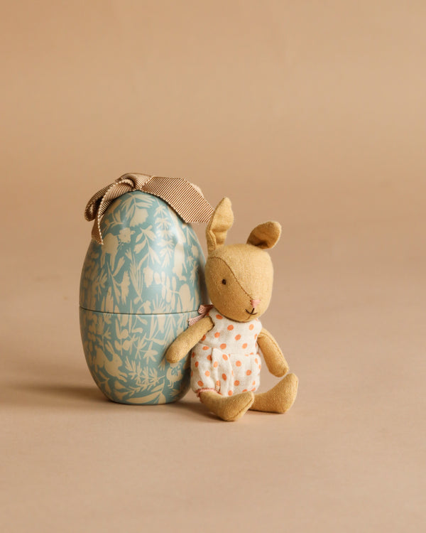 A Maileg Easter Egg in dusty blue with a floral pattern sits on a beige background beside a soft micro rabbit gift in a white dress with orange dots.