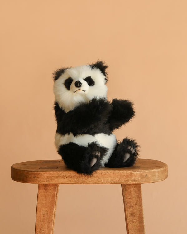 The Panda Cub Stuffed Animal, featuring soft black and white fur, sits upright on a wooden stool with a plain beige background.