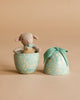 The Maileg Easter Egg with Micro Lamb, Small - Dusty Mint, features a plush magnetic lamb with a green bow inside a decorative light blue floral egg-shaped container. The matching lid, also topped with a green bow, rests beside it.