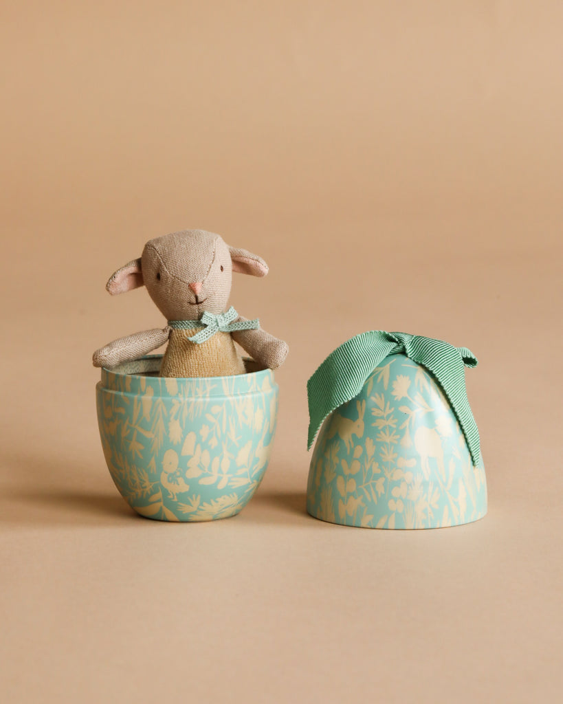 The Maileg Easter Egg with Micro Lamb, Small - Dusty Mint, features a plush magnetic lamb with a green bow inside a decorative light blue floral egg-shaped container. The matching lid, also topped with a green bow, rests beside it.