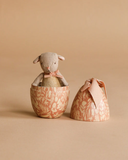 The Maileg Easter Egg with Micro Lamb, Small - Powder features a soft lamb toy with a pink ribbon inside a floral-patterned metal egg. The matching ribbon-tied lid rests nearby—an adorable children’s gift.
