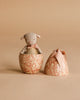 A small stuffed animal from the Maileg Easter Egg & Friends Set peeks out of a decorative beige egg with a floral pattern, while the other half featuring a bow sits next to it on a matching beige background.