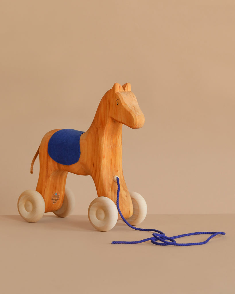 Grimm's Horse Holly, a charming wooden pull along toy with white wheels, a blue string, and a blue saddle, rests on a neutral beige surface against a soft beige background.