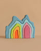 Pastel-colored wooden arches from Grimm's Blue Cave Arch Stacking Toy, resembling abstract mountains, stacked on a plain beige background. The arches come in blue, green, yellow, orange, pink, and purple.